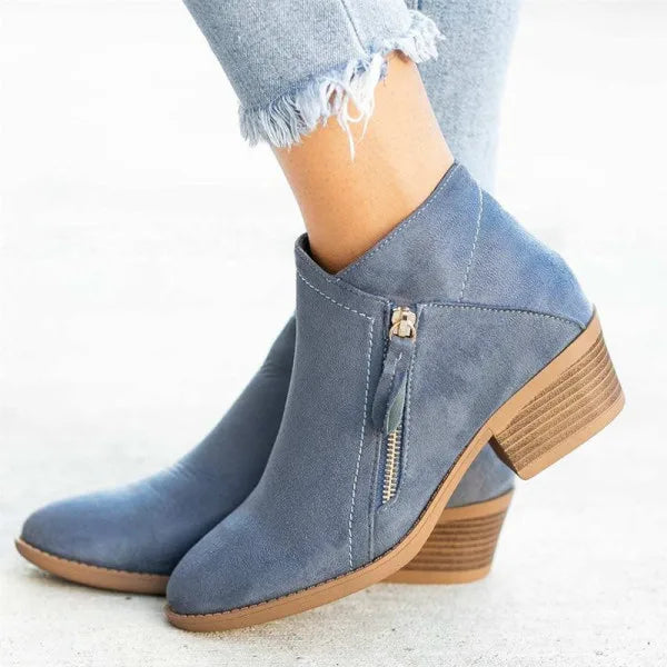 Side view of blue suede ankle boots with a stitched V-seam, gold zip fastening and stacked block heel, worn with frayed denim jeans.