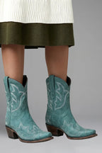 Turquoise teal suede ankle boots with white embroidered stitching worn beneath a knit skirt hem