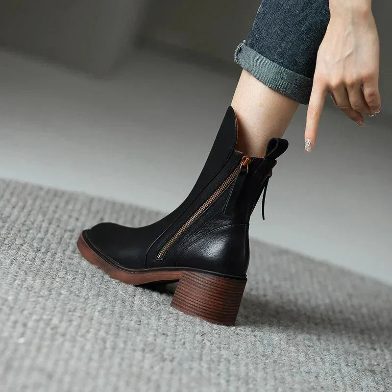 Close-up of black leather ankle boot on model's foot with gold-tone zip and pull tab, on grey carpet