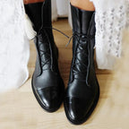 Front view of black leather lace-up ankle boots with cap toe, worn with a white lace hem dress on wooden floor