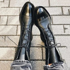 Top-down view of black lace-up boots showing crossed laces and glossy cap toe detail against paving stones