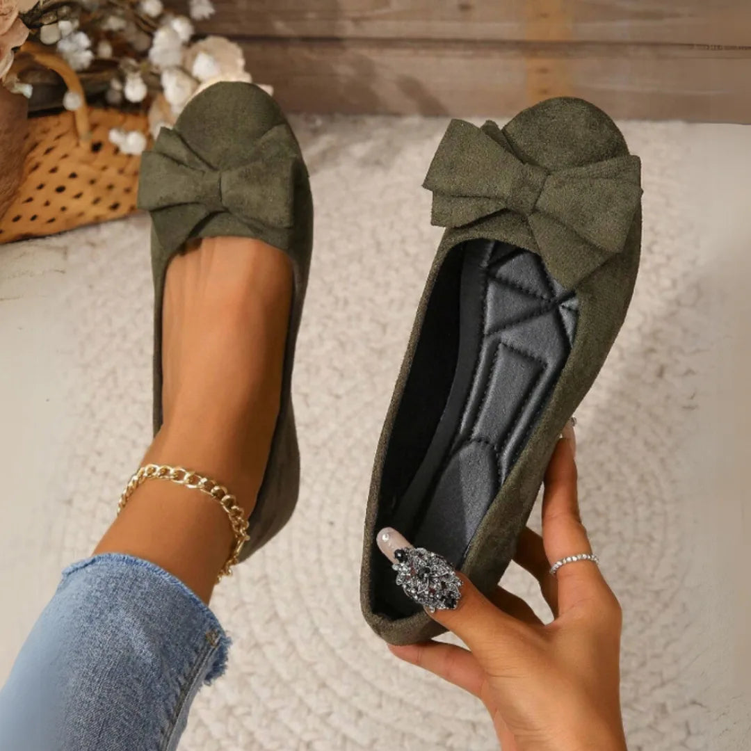 Dark green suede-effect bow ballet flats shown on foot next to a hand holding the second shoe, revealing the padded grey insole
