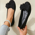 Black suede-effect ballet flats with bow detail, one worn and one held to show the cushioned quilted insole