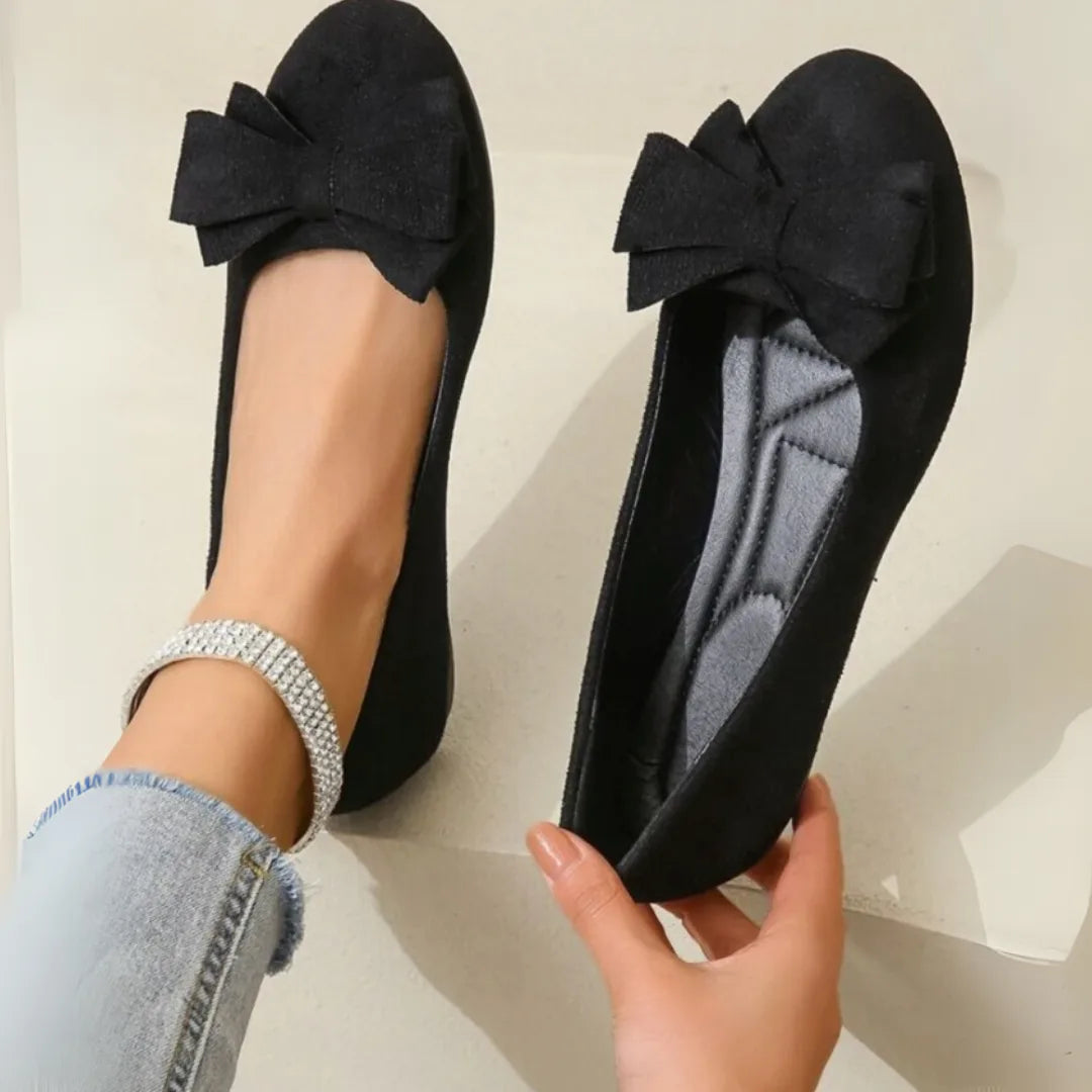 Black suede-effect ballet flats with bow detail, one worn and one held to show the cushioned quilted insole