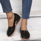 Black perforated leather flat with elasticated strap, paired with denim trousers