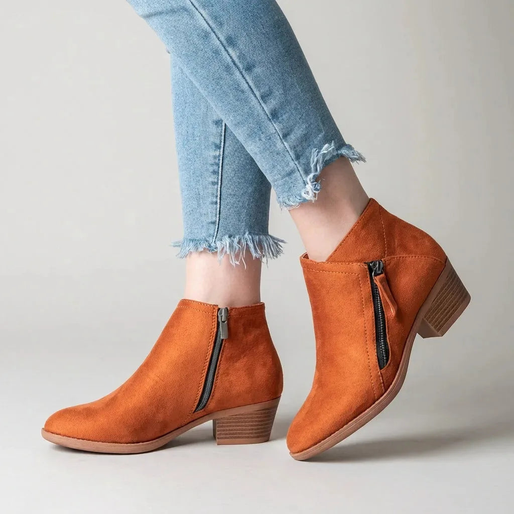 Orange faux-suede ankle boot with contrast zip pull, worn with cropped jeans against a neutral backdrop