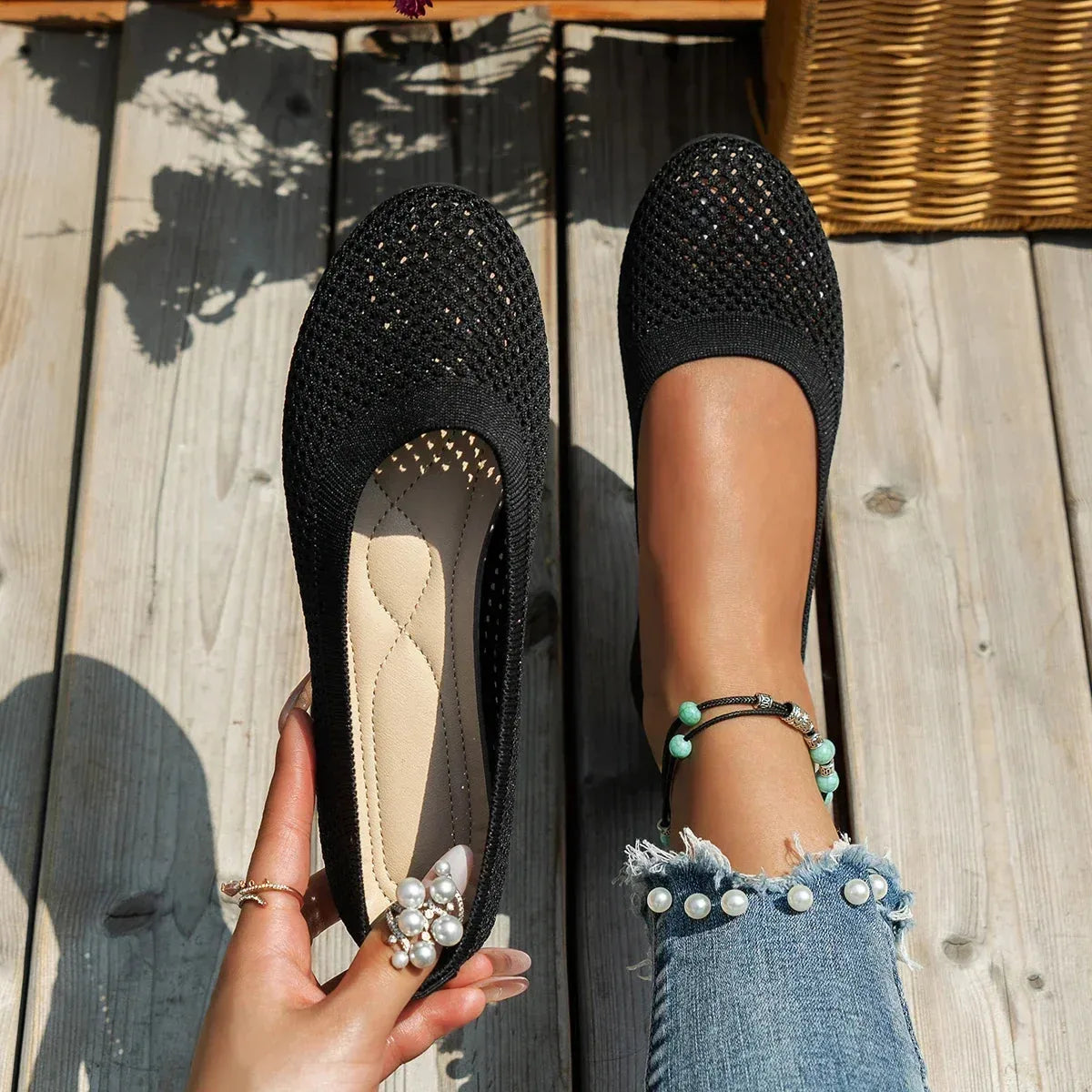 Black knit mesh ballet flats shown from above, held by hand and worn on foot on wooden decking