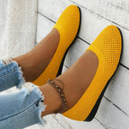 Yellow ballet flat held against wooden background, showing flexible knit sole and stitched trim