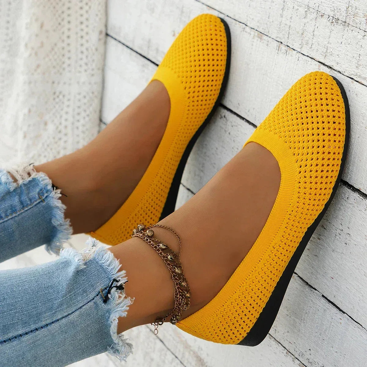 Yellow ballet flat held against wooden background, showing flexible knit sole and stitched trim