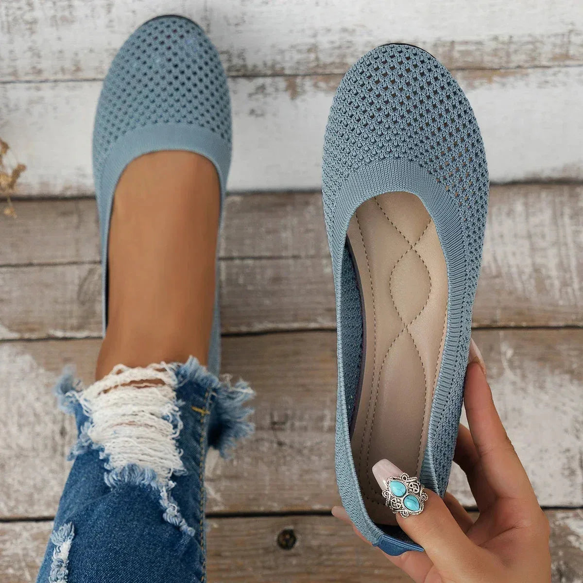 Grey-blue knit ballet flats shown from above on distressed wooden decking, one held by hand