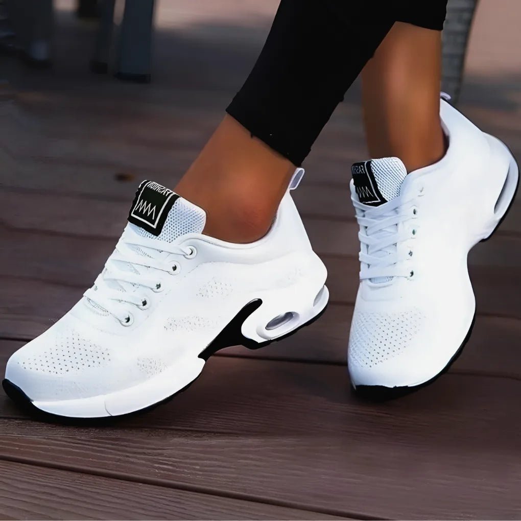 Angled view of white mesh trainers with black sole accents and dark heel tab, worn on a wooden deck