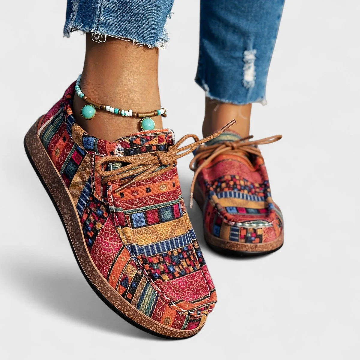 Close-up front view of red and orange patchwork lace-up comfort shoes worn with cropped jeans and a beaded anklet.