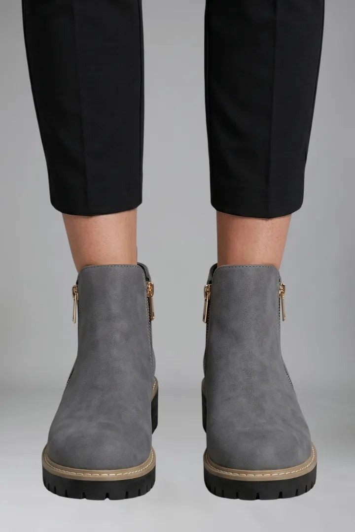 Close-up front view of grey ankle boots showing gold zip pulls and stitched tan trim on a black lug sole