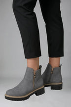 Side view of grey suede-effect ankle boots worn with cropped black trousers, showing the zip and heel pull tab