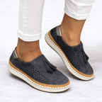 Charcoal grey suede slip-on trainers worn on feet, showing metallic heel tab and stitched sole detail