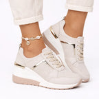 Front angle of beige wedge trainers with gold trim, perforated toe panel and lace-up fastening