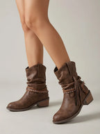 Side view of brown slouch ankle boots with braided wrap straps and leather tassel worn on model's calves