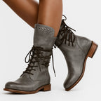 Side view of grey leather lace-up mid-calf boots worn on crossed legs, showing studded cuff and laces