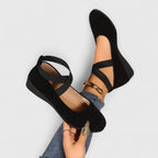 Black suede ballet flats with crisscross ankle straps and a low wedge sole, shown on model's raised feet
