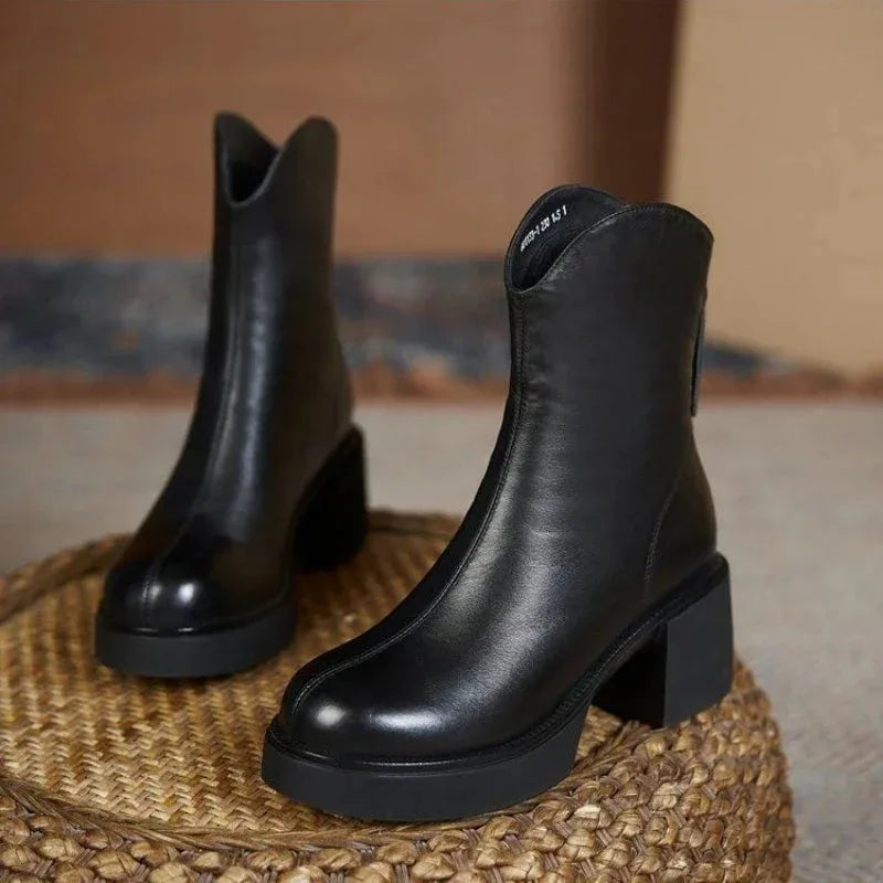 Pair of black leather Chelsea boots resting on a woven rattan stool, showing smooth finish and stacked heel