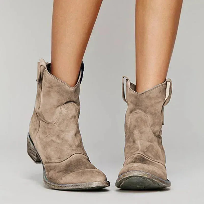 Front view of tan suede cowboy ankle boots with distressed finish and pointed toe, worn on bare legs.