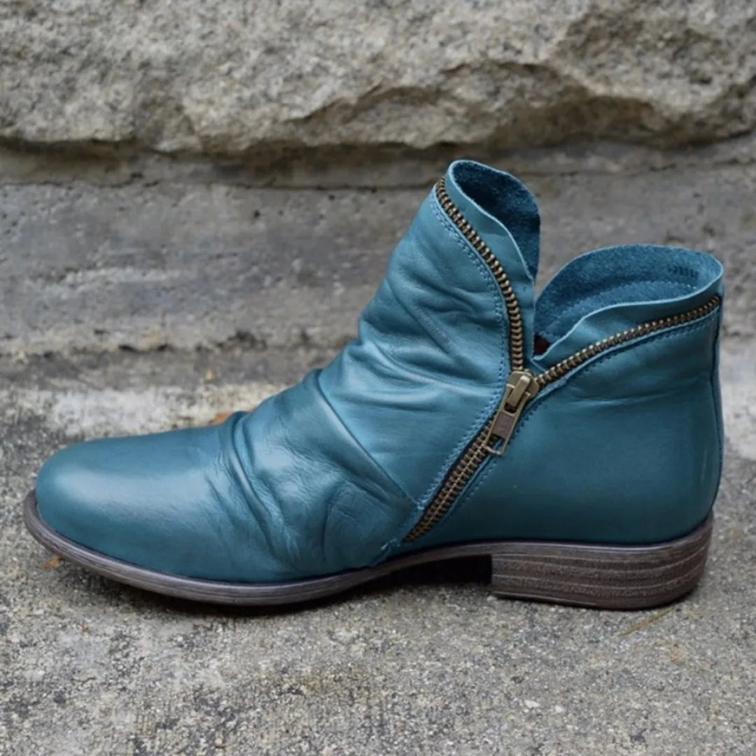 Teal leather ankle boot with bronze-tone zip and soft slouched shaft, photographed against a stone wall.