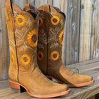 Pair of tan leather cowboy boots with hand-stitched sunflower and leaf embroidery on the shaft, shown against a wooden fence.