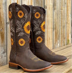 Dark brown leather cowboy boots with orange and yellow sunflower embroidery, standing on a wooden deck.