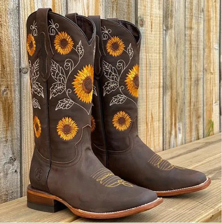 Dark brown leather cowboy boots with orange and yellow sunflower embroidery, standing on a wooden deck.
