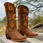 Brown leather cowboy boots with sunflower stitching detail, photographed outdoors on a stone ledge with greenery behind.