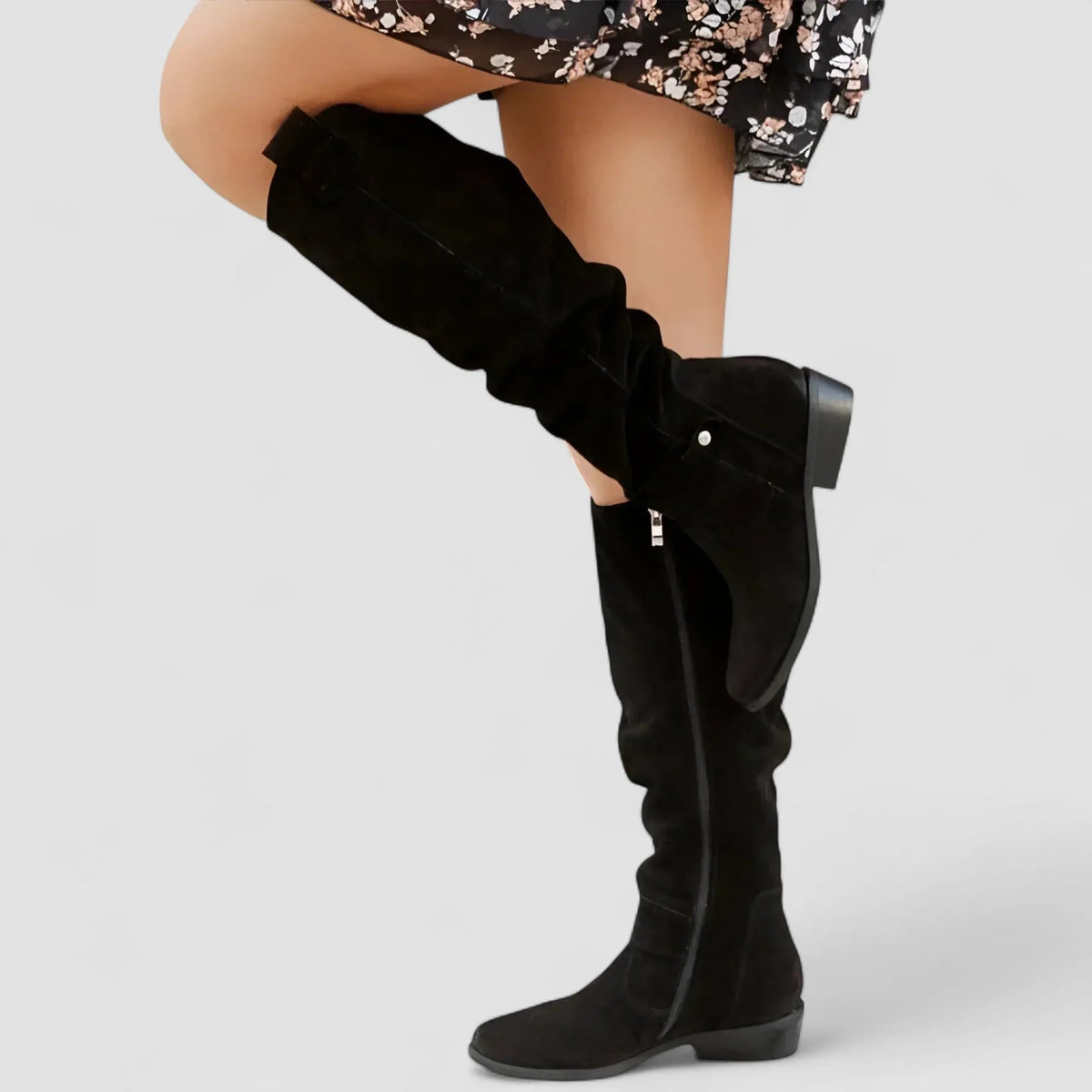 Black suede knee-high slouch boot with side zipper and pull tab worn under a floral dress