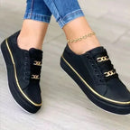 Black faux-leather lace-up trainers with gold chain detail and gold piping, worn on feet with cropped jeans