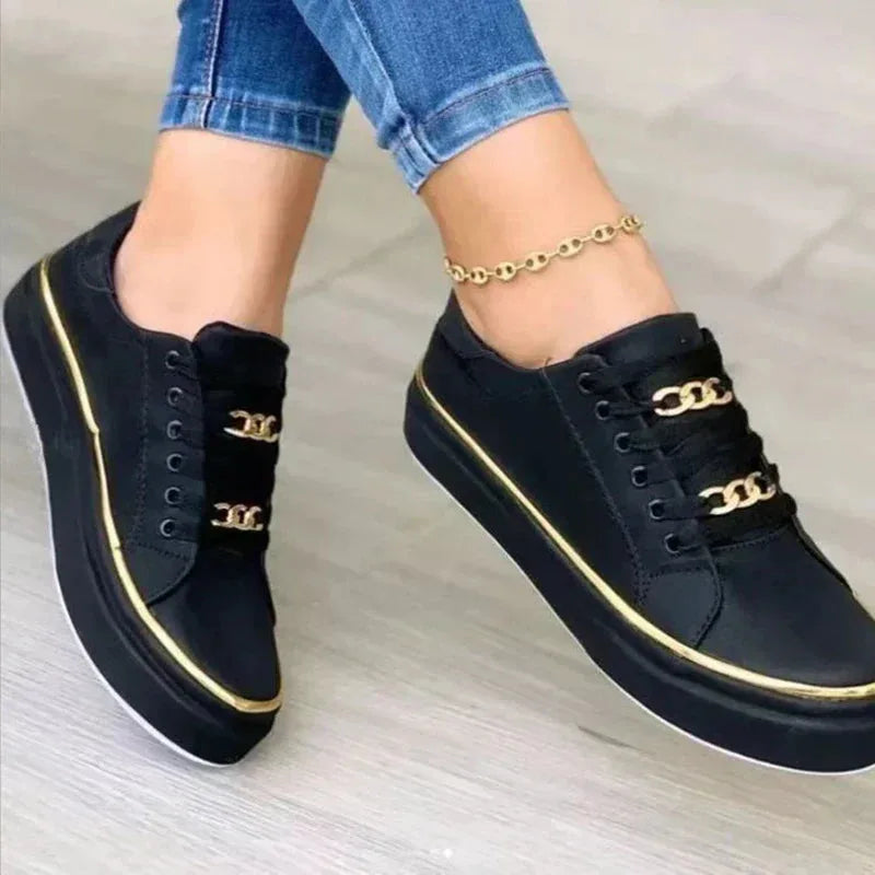 Black faux-leather lace-up trainers with gold chain detail and gold piping, worn on feet with cropped jeans