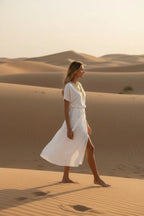 Woman walking barefoot across sand dunes in the flowing ivory wrap midi dress, side view.