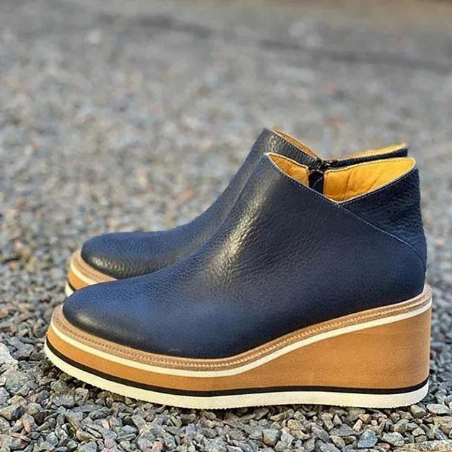 Crossed-ankle view of tan-brown leather wedge boots on wooden floor showing pointed toe shape